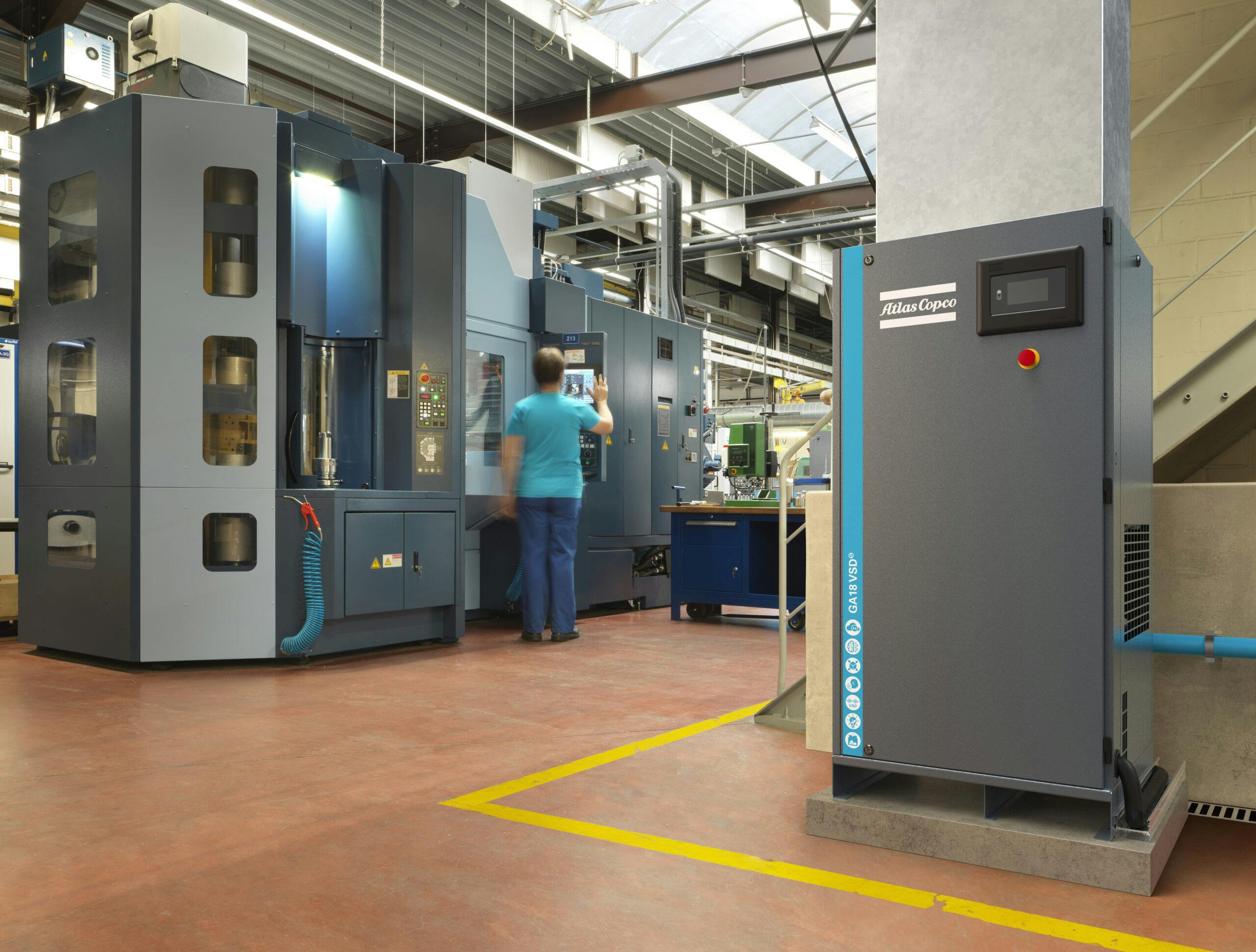 Atlas Copco Kompressor i produksjonshall, medarbeider jobber med produksjon i CNC maskin. Kompressor er koblet til airnet rør.